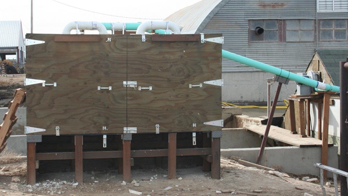A raised plywood cabinet outdoors has PVC pipes going into it.